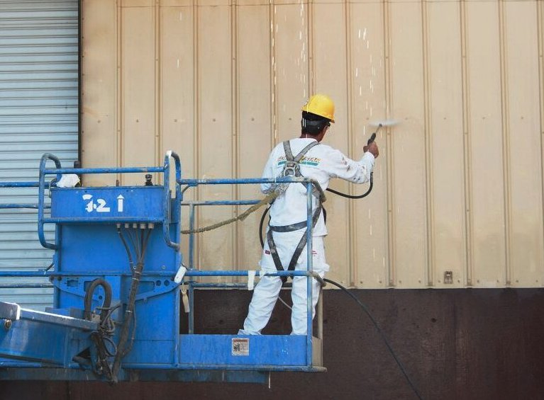 Worker on a lift high-pressure washing a metal building wall before coating