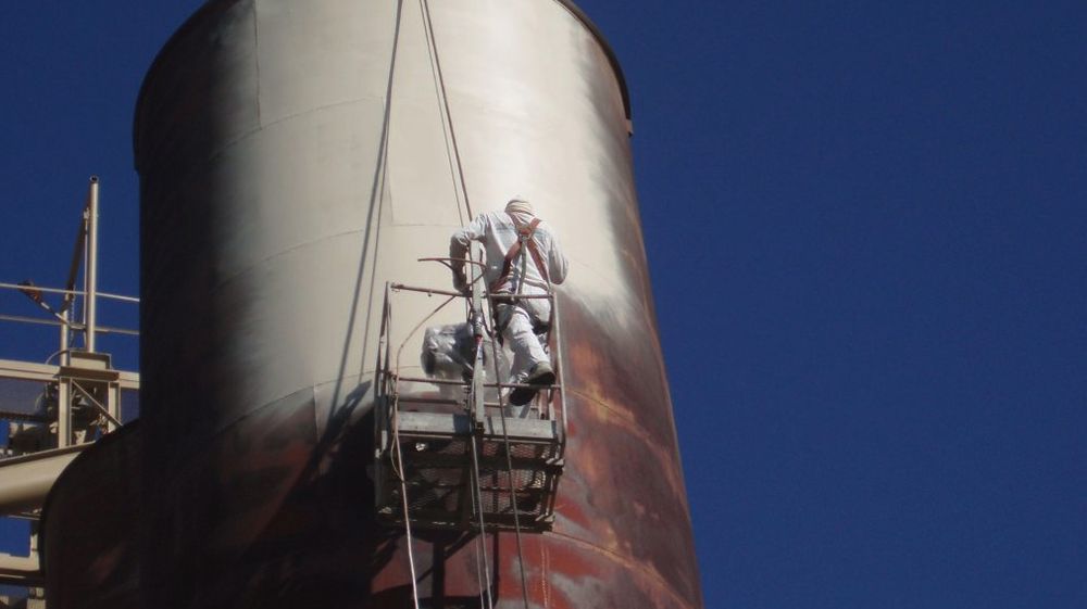 Gold Star Painting crew member on a suspended platform spray-coating a large industrial storage tank