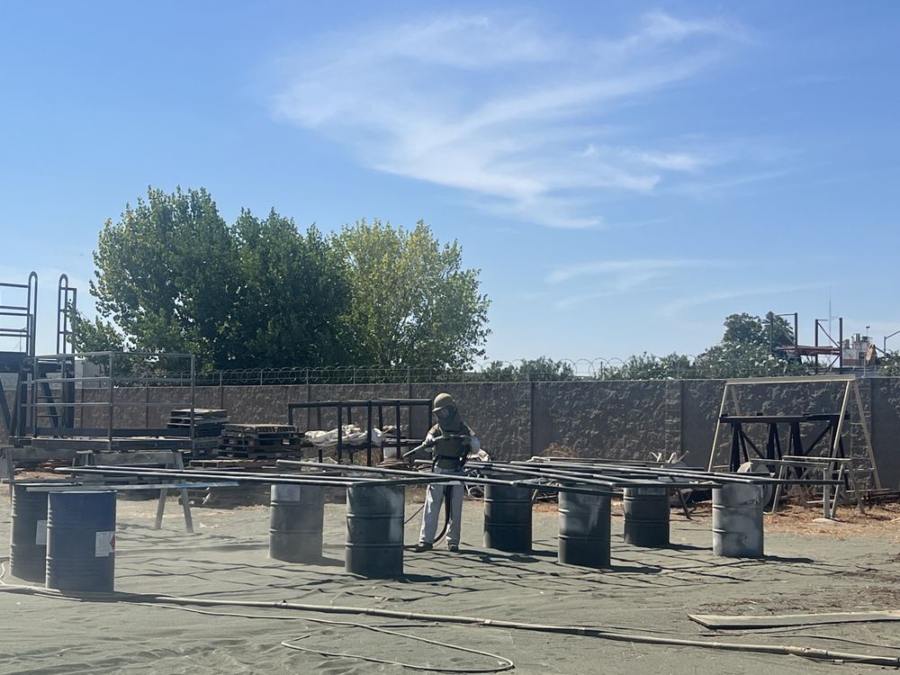 Gold Star Painting technician in a blast hood sandblasting steel railing sections at the Modesto yard